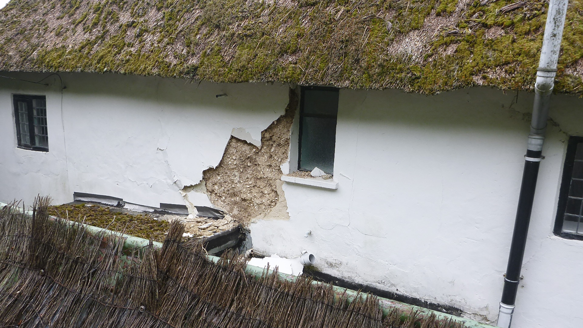 before photo of render falling down on side of white house with thatched roof