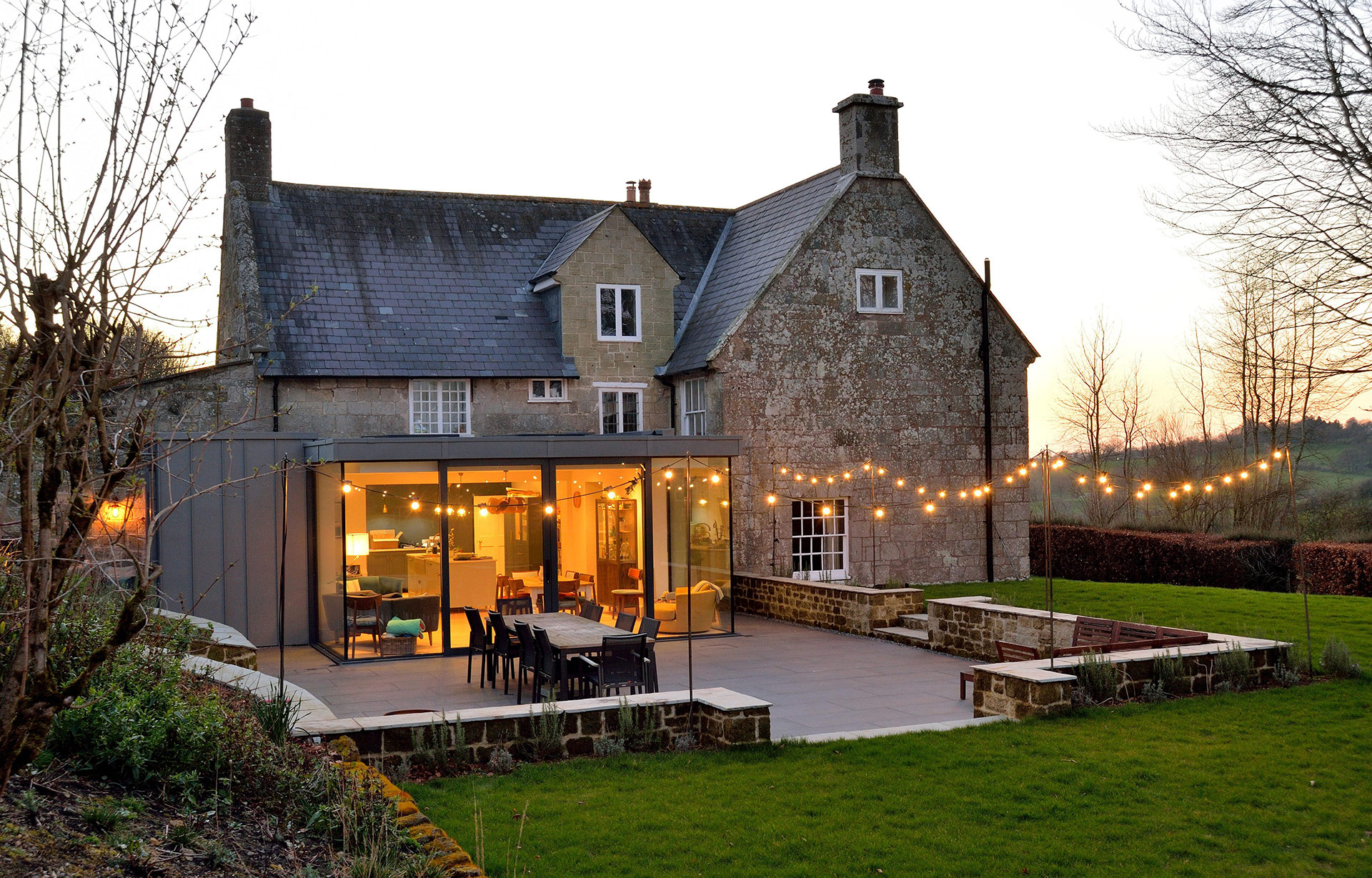 modern extension of traditional stone house with large patio area taken at dusk with lights on