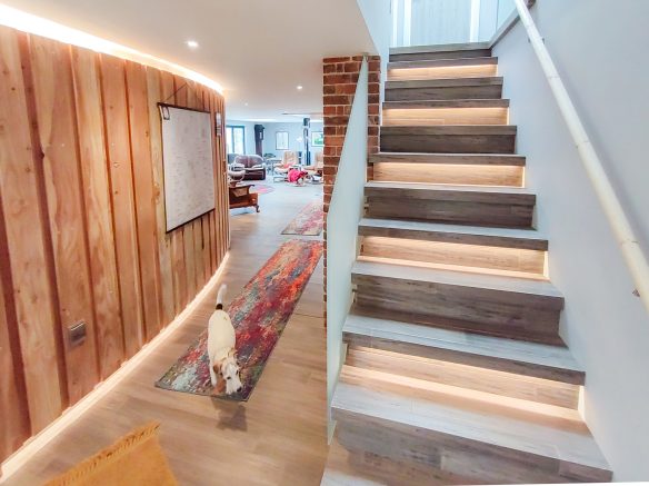 wooden stairs with lights under step leading from open plan living area with curved timber clad wall